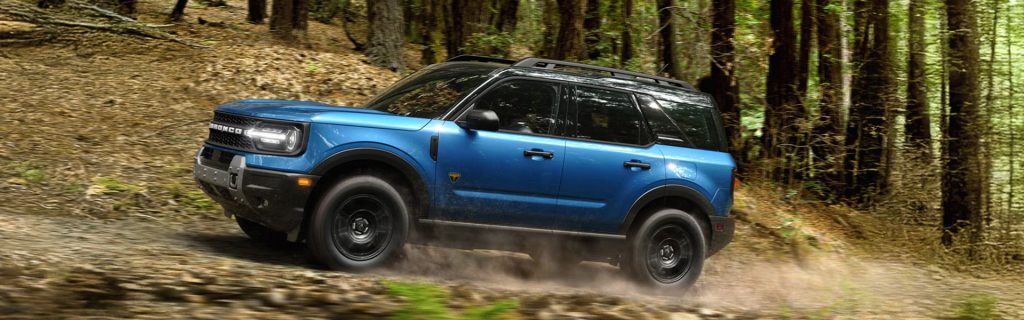 Blue Ford Bronco driving off-road through a forested area.