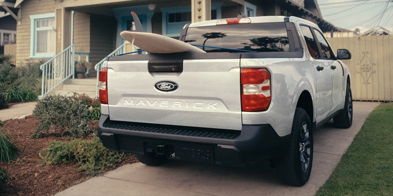 Rear view of a white Ford Maverick pickup with a surfboard in the bed parked in a driveway.